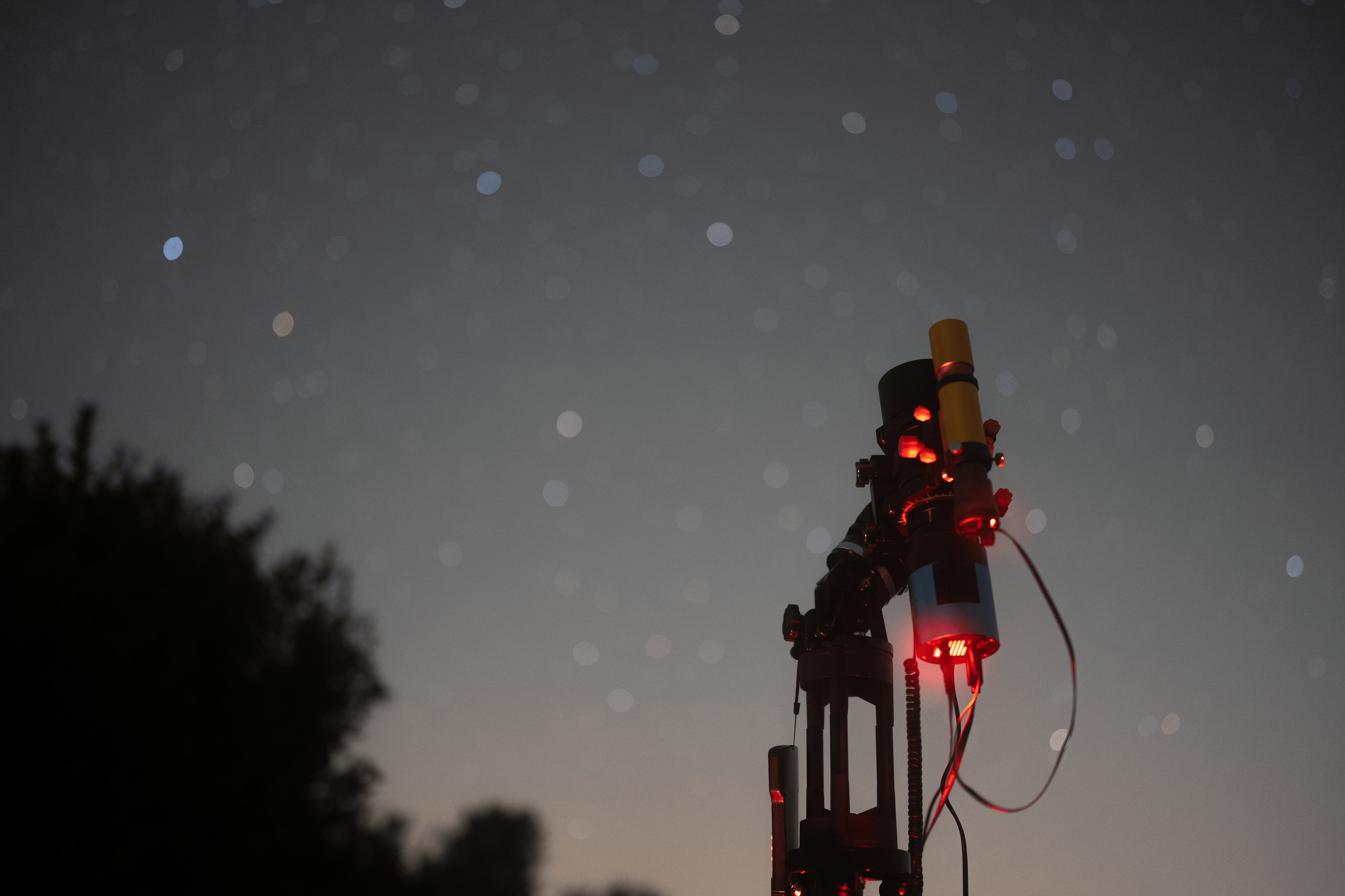 A second nightscape image of the telescope setup from a different angle. The scope is pointing towards the sky full of stars. In the back a tree can be seen.