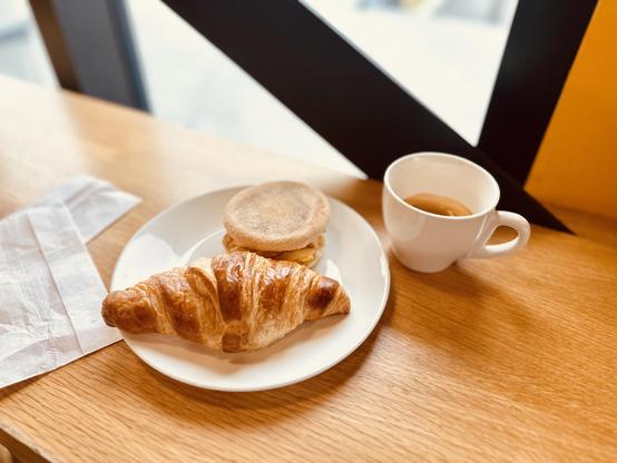Ein kleiner Teller mit einem goldbraunen Croissant und einem Muffin. Daneben eine grosse Tasse mit Kaffee.