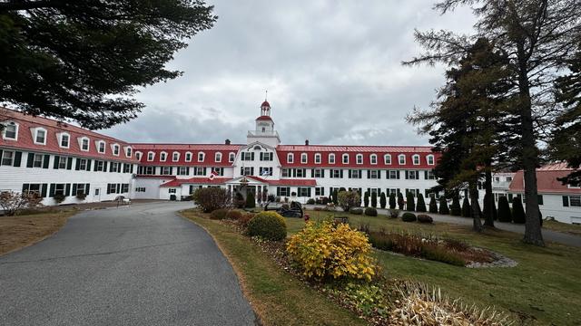 Hotelkomplex mit weißer Basis und roten Obergeschoss. L-förmig, in der Mitte die Reception, welche ein wenig an einen Leuchtturm gemahnt. Rundherum ein paar Bäume und ein Rasen mit Sträuchern und Blumen. Der Himmel ist grau bedeckt.