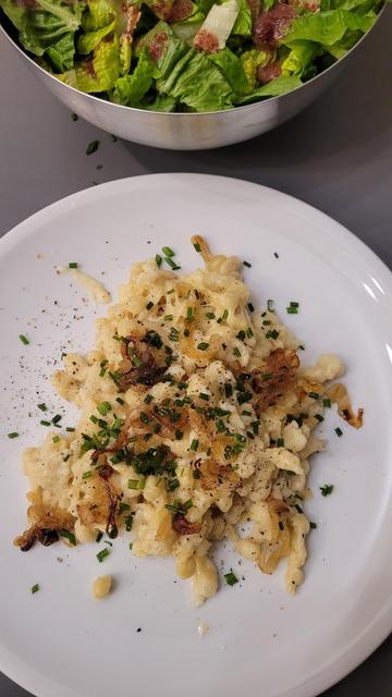 Farbfoto: Eine Portion Käsespätzle mit Röstzwiebeln und Schnittlauch, über dem Teller ist (angeschnitten) eine Schüssel mit Salat mit rötlichem Dressing.
