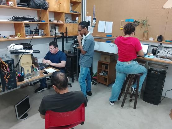 2 pessoas pessoas sentadas e 1 em  pé ao redor de um computador instalando linux. Uma pessoa de costas apoiada em um banco instalando Linux em um notebook em uma bancada.