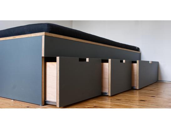 Natürlich! Hier ist eine detaillierte Beschreibung des Bildes für Menschen mit Sehschwäche:
Das Bild zeigt ein modernes, hoch gebautes Bett oder eine Sitzbank mit Stauraum darunter. Das Möbelstück ist aus Holz und hat eine klare, geradlinige Form.

Oberfläche: Auf der Oberseite liegt eine dunkle, gepolsterte Matratze oder Auflage, die sich über die gesamte Länge erstreckt.
Seitenteile: Die Seiten des Möbels sind mit dunklen, glatten Platten verkleidet, die in einem tiefen Grau oder Anthrazit gehalten sind.
Front: Die Front besteht aus mehreren Schubladen oder Klappen, die ebenfalls dunkelgrau sind. Diese haben kleine, horizontale Griffmulden, die in die Oberfläche eingelassen sind, um das Öffnen zu erleichtern.
Holzrahmen: Der Rahmen des Möbels ist aus hellem Holz gefertigt, das an den Kanten und als vertikale Streben zwischen den dunklen Platten sichtbar ist.
Boden: Das Möbel steht auf einem hellen Holzfußboden, der eine natürliche, warme Farbe hat.
Wand: Im Hintergrund ist eine weiße Wand zu sehen, die dem Möbelstück einen klaren Kontrast bietet.

Das Möbel wirkt funktional und stilvoll, mit einer Kombination aus dunklen und hellen Elementen. Die Struktur ist stabil und bietet durch die Schubladen oder Klappen Stauraum.