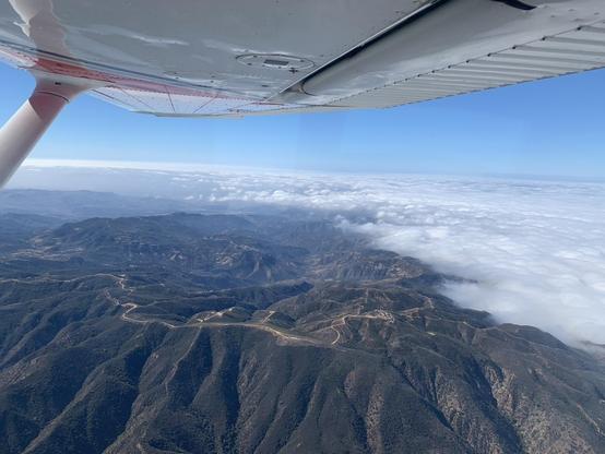 Aerial view of a layer of cloud, ending at a mountain range. They are seen from above, with blue skies and the wing of the airplane seen at the top of the image