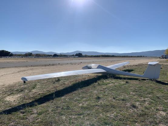 A slender, white glider sits tied down in the grass beside a runway on a sunny day