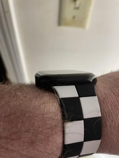 In the foreground, a caucasian man's left forearm displaying an Apple Watch with a checkered black and white band that resembles the classic black and white checkered pattern associated with the music.