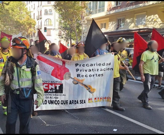 Fotografía de una manifestación de bomberxs forestales y trabajadorxs con camisetas amarillas, cascos y banderas rojinegras. En el centro se sostiene una pancarta con el logo de CNT-AIT y el mensaje: “Precariedad, Privatización, Recortes. Que lo único que arda sea el capital”.