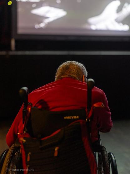 An elderly person in a wheelchair is seen from behind, wearing a red jacket. They are watching a performance on a screen, which shows a black-and-white image of a guitar. The setting appears to be a dimly lit venue.
