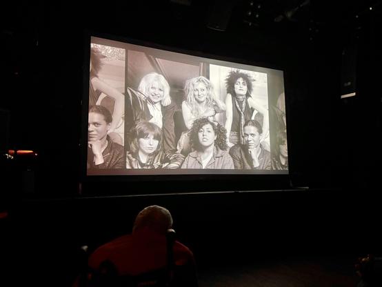 A black-and-white photograph projected on a screen features a group of diverse individuals posing together. The scene appears to be from a past era, showcasing various hairstyles and fashion styles. In the foreground, a person seated in a wheelchair is partially visible.