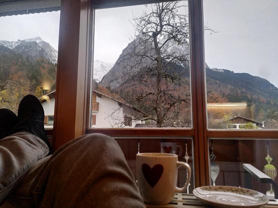 Ein Blick aus einem Balkonfenster auf eine herbstliche Alpenlandschaft. Auf einem Heizkörper liegen meine Beine ind eine Tasse Kaffee habe ich dort abgestellt.