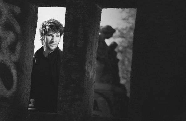 Damian Scholl with a thoughtful expression is framed by a stone structure in a black and white photo, with blurred trees and a statue in the background.