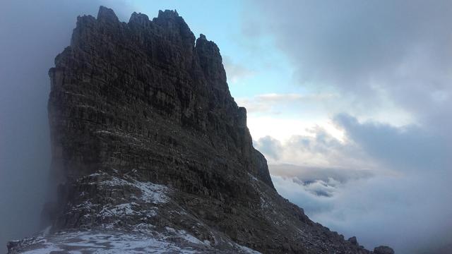 Eine Felsengruppe mit leichtem Schnee auf dem Boden davor. Der Nebel zieht hoch während rechts noch ein bisschen blauer Himmel zu sehen ist und auch dräuende Wolken.