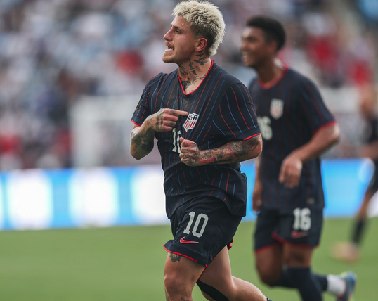 Soccer player with short grey hair has heavily tattoed arms and neck wearing a black shirt with red and lines,  is pointing at a US Men's National Team shield on his chest and has the number 10 and a Nike swoosh on his shorts.  Number 16, a black player is behind him and out of focus and past that are blurry bleachers and a video sign with white almost rectangular puffs on a blue background