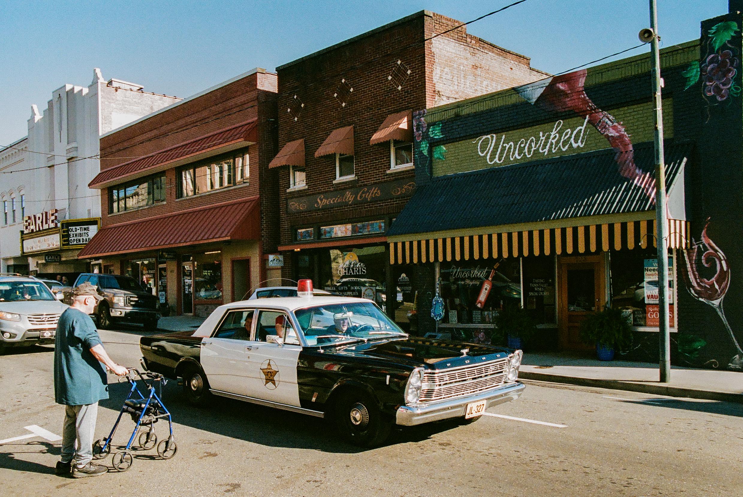 A photo of early 1960s Ford police car driving on the street of a small town.  There is an older guy with a rolling walker/buggy waiting to cross the street.