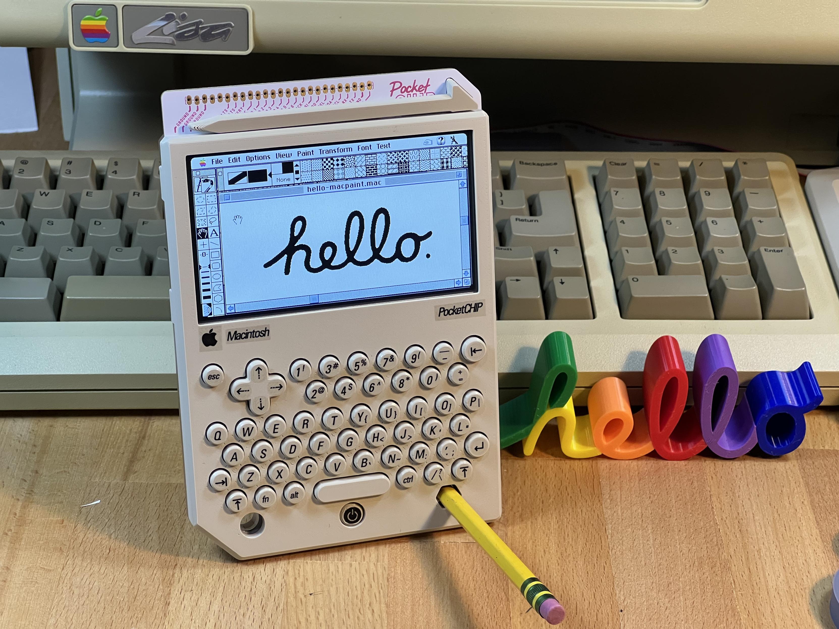 A PocketCHIP hand held computer on a desk using a #2 pencil as a stand. The case is in a warm beige color with round keys. On the screen is Mac System 7.5 with SuperPaint loaded. A document is open showing cursive hello. On the desk is a rainbow 3D printed cursive hello in front of a Lisa computer and keyboard.