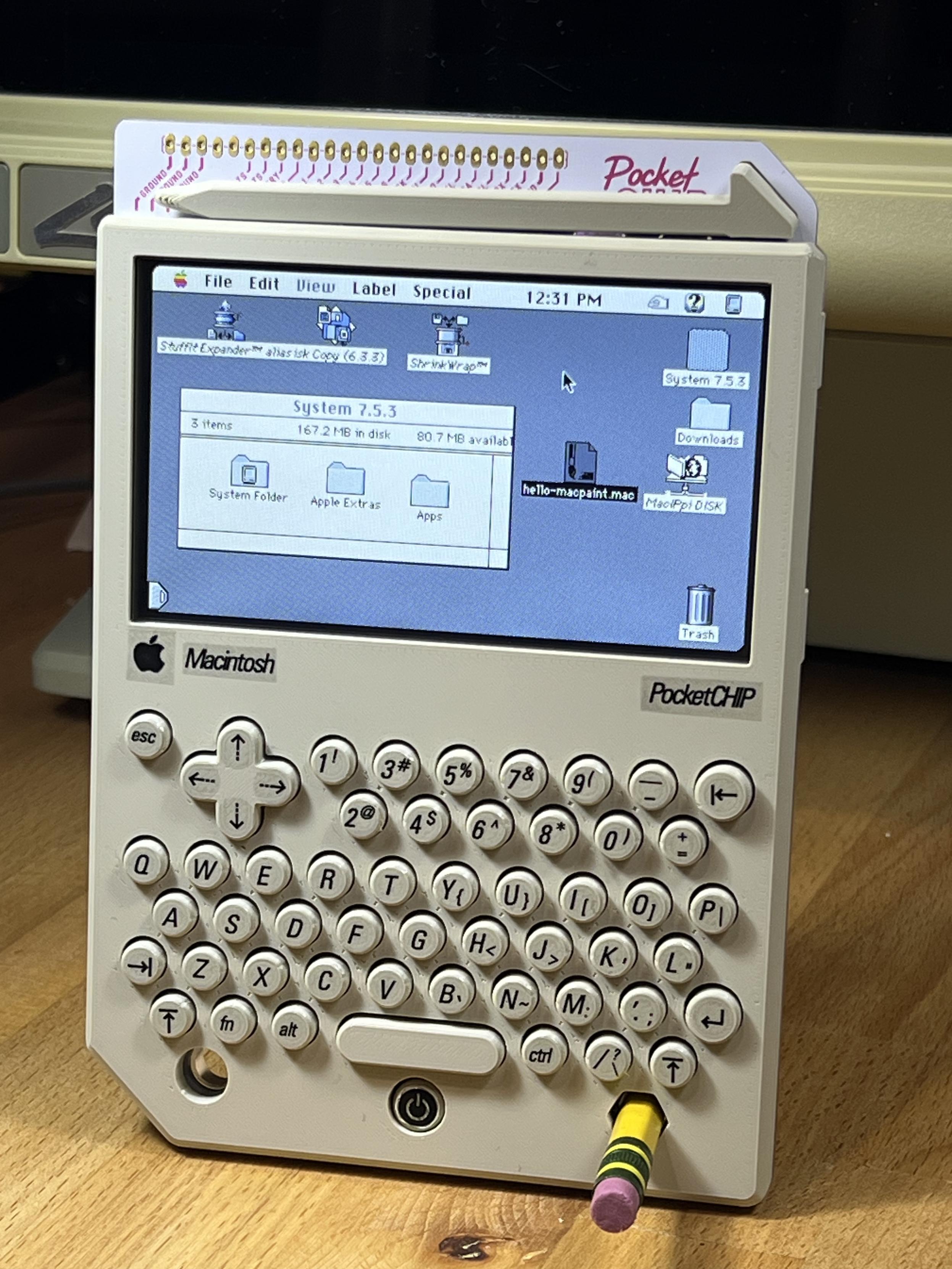 A PocketCHIP hand held computer on a desk using a #2 pencil as a stand. The case is in a warm beige color with round keys. On the screen is Mac System 7.5 with a window open and some icons on the desktop.