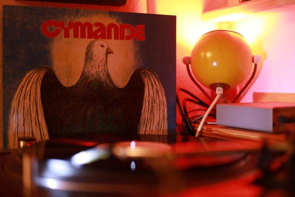 In the foreground, a blurred record player; in the background, the album cover of Cymande Cymande next to a round yellow lamp.