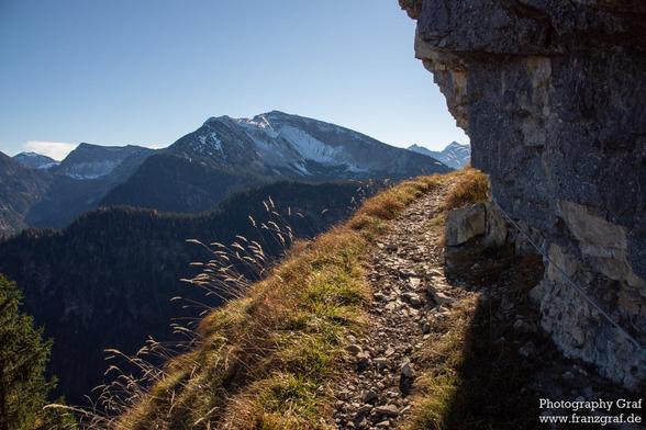 A rugged stone path winds gracefully along the edge of a steep, sunlit slope, inviting hikers deeper into the high alpine wilderness. Golden grasses sway gently in the breeze, their warm hues contrasting beautifully with the cool, shadowed valleys below. Towering above, a majestic peak—dusted with patches of lingering snow—dominates the skyline, its sharp ridges cutting crisply against the deep blue sky. The air feels clear and expansive, with layer upon layer of forested hills rolling into the…