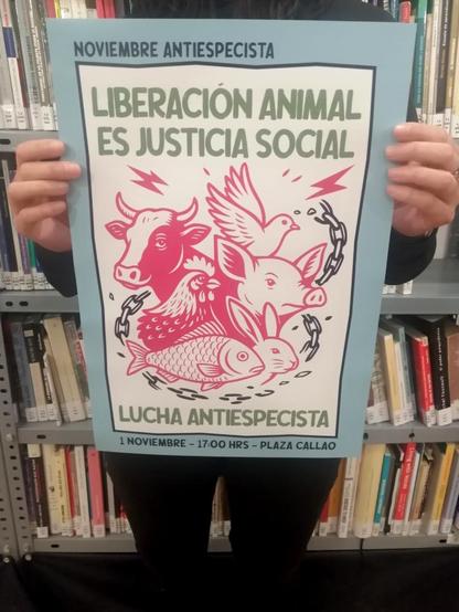 Foto de una mujer sujentando un cartel de la manfiestación antiespecista del 1N en Madrid. A las 17:00 en Callao, con el lema "Liberación animal es justicia social. Lucha antiespecista". No se ve el rostro de la mujer y de fondo hay unas estanterías con libros, vamos, que está en la biblioteca del Local anarquista Magdalena.