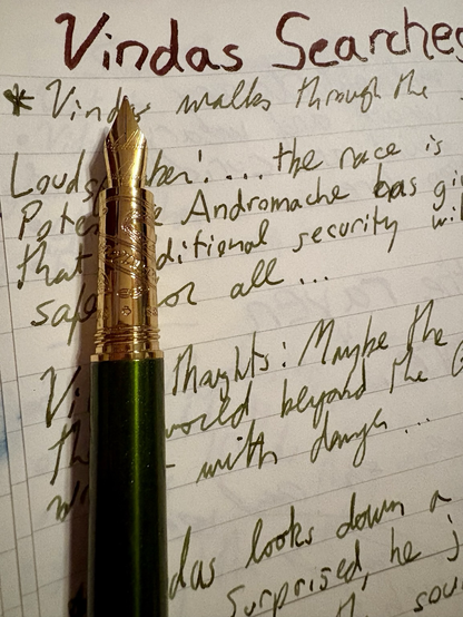 A green fountain pen with etched gold at the nib and section, rests upon a page with notes. The top of the page is labelled “Vindas Searches” in thick, dark red letters. The pen covers the writing I part, and the photo cuts off the words. Names like Vindas and Andromache stand out… it speaks about a race and of security, and that maybe the world beyond is wrought with danger…