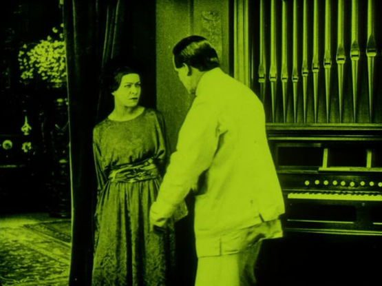 an image of a man and woman in front of an organ, the woman looking at the man scornfully, an open door behind her, the film is tinted yellow