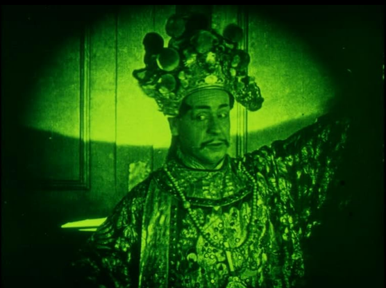 an image of a man with an ornate chinese headdress on, raising one arm in the air, looking to his left, the film tinted green