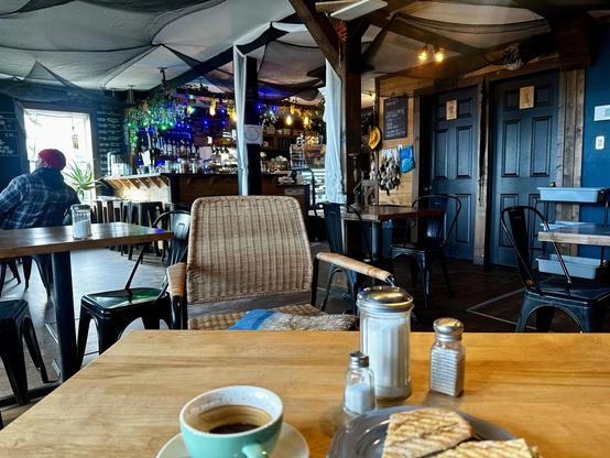 Szene im Café. Vor mir auf dem Tischchen ein doppelter Espresso in einer grünen Tasse. Daneben ein grilliertes Sandwich.
Das Café ist dekoriert mit viel Nippes.