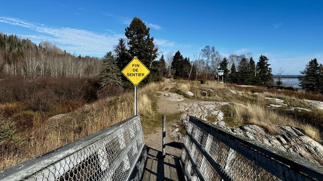 Blick von einer schmalen Holzbrücke auf das Ende. Gelbes Warnschild: «Fin du Sentier».
Dahinter, ein steiniger, sandiger Weg und Bäume.