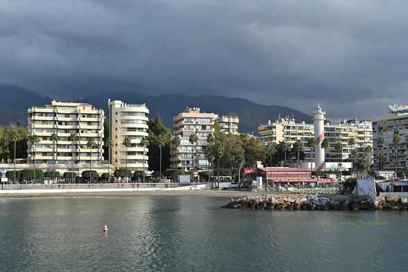 Imagen horizontal donde se ven edificios y el faro de la ciudad a mediación del cuadro. El tercio superior es cielo muy nublado, que amenaza lluvia, y el tercio inferior es mar.