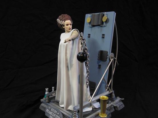 X-Plus bride of Frankenstein model kit. Gowned Bride with tan bandages. On a stone base with various metal work and scientific equipment. A black background.
