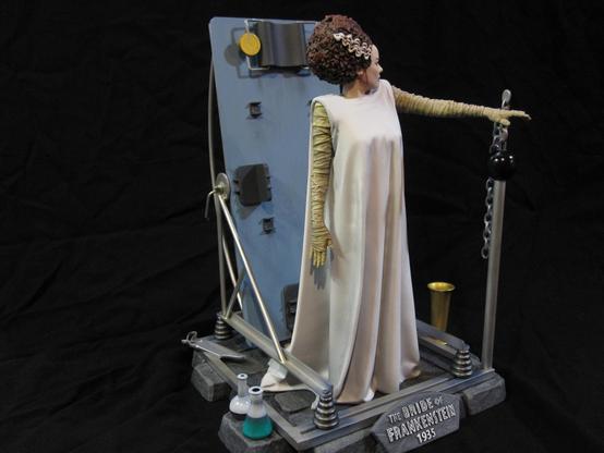 X-Plus bride of Frankenstein model kit. Gowned Bride with tan bandages. On a stone base with various metal work and scientific equipment. A black background.