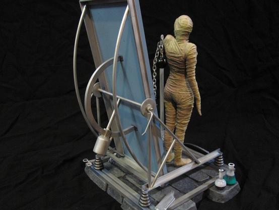X-Plus bride of Frankenstein model kit. Tan-bandaged Bride. On a stone base with various metal work and scientific equipment. A black background. Rear view.