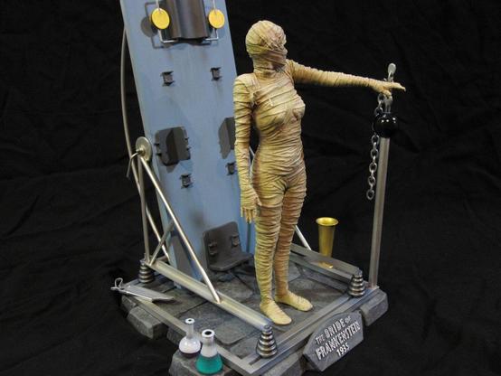 X-Plus bride of Frankenstein model kit. Tan-bandaged Bride. On a stone base with various metal work and scientific equipment. A black background.