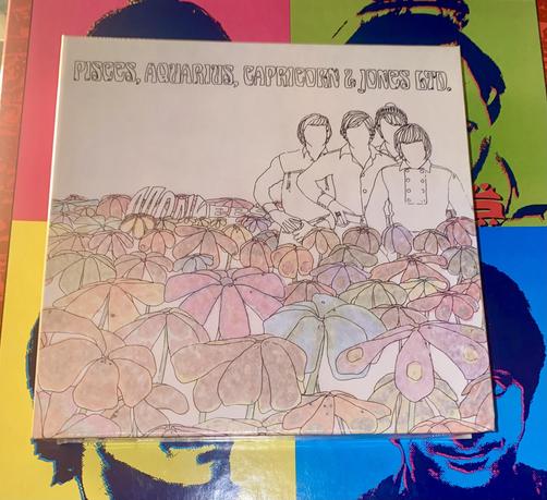 The image features a CD box cover titled "Pisces, Aquarius, Capricorn & Jones Ltd." by the Monkees. It showcases a line drawing of four figures surrounded by colorful flowers in shades of pink, blue, and green. The background includes