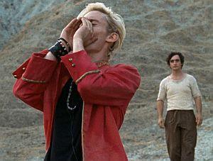 Zwei junge Männer stehen in einer öden Landschaft. Ein Mann, gekleidet in einer roten Jacke und mit Armschmuck und Halskette, hält die Hände neben den Mund um seine Nachricht zu verstärken. Der zweite Mann, ärmlicher gekleidet, sieht im zu.