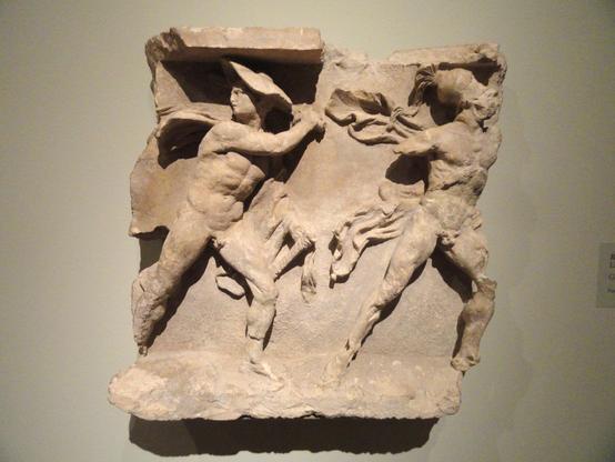 This carved limestone relief was likely part of a larger frieze that decorated a naiskos, or small temple, above a grave.
The relief depicts two warriors striding to the right, one wearing a broad traveller's hat called a petasos and the other a Corinthian helmet. Each wears a short cloak called a chlamys over his left arm (now missing on the righthand warrior). The left warrior looks back, perhaps toward a horseman, since a small fragment of a horse’s foreleg remains behind his knee. The stron…