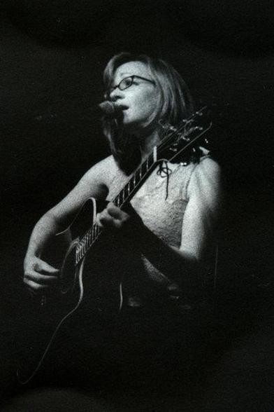 Lisa Cerbone plays an acoustic guitar and sings into a microphone on a dimly lit stage. She wears glasses and a sleeveless top, evoking a serene mood in this low quality image.