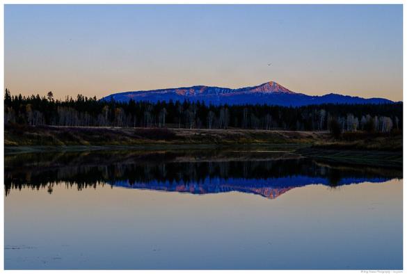 A snowcapped mountain, tinged by sunset light, is seen in the distance above a forest and grassy shoreline, with its reflection mirrored in the calm surface of a river in the foreground.