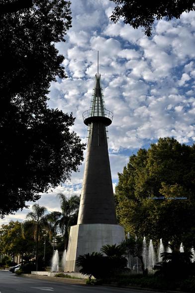 Imagen de un monumento en forma de cono situado en una glorieta de unos veinte o treinta metros de altura. Está sobre una base blanca, parece que de mármol, y la mayor parte del cono es metálica, posiblemente cobre. El vértice del cono es de cristal. La imagen también muestra ramas de algunos árboles del lugar y cielo con nubes el forma de algodones.