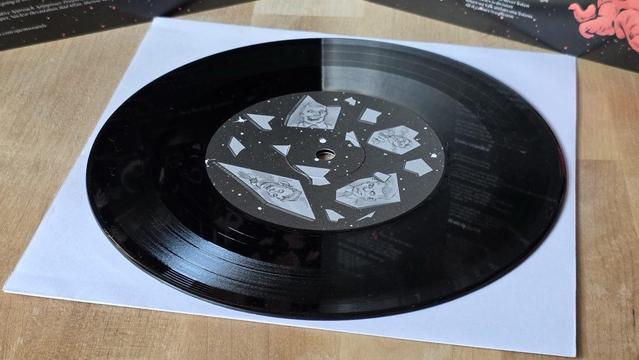 Eine künstlerische schwarze Schallplatte auf einer weißen Papierhülle. Das Label hat einen dunklen Hintergrund mit Illustrationen von Gesichtern und geometrischen Formen, die an ein Design mit kosmischem Thema erinnern.