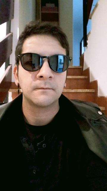 Señor moreno(yo) con gafas de sol, pendiente con una pluma negra colgandera, camisa negra con el botón abierto y una chaqueta de polipiel a los hombros.