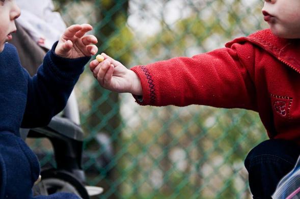 Share information don't hoard it: A photograph of two young children, one in a red coat, one in blue. The former is offering a yellow candy to the latter; their hands are about to touch.
