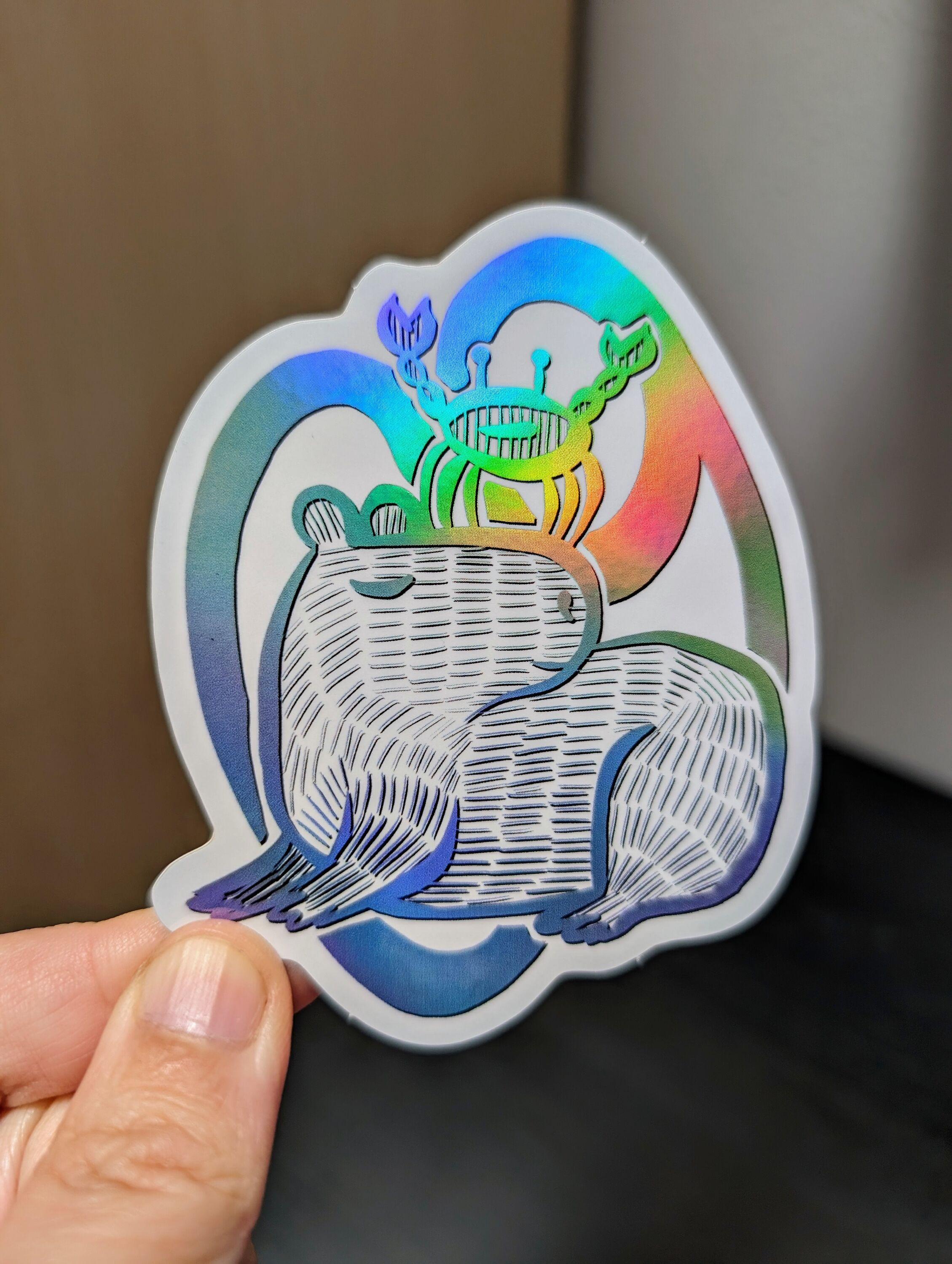 Photo of a left hand holding a diecut sticker. The sticker is a holographic drawing of a capybara Cancer zodiac sign.