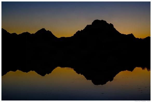 A silhouetted mountain range is reflected in calm water below, with a gradient sky transitioning from deep blue at the top to golden yellow along the horizon, suggesting sunrise or sunset.