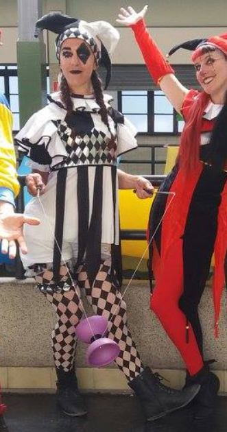 Fotografía donde se ve a dos mujeres, la de la izquierda vestida de arlequin en blanco y negro, con un diábolo morado. La derecha, vestida de bufón en rojo y negro