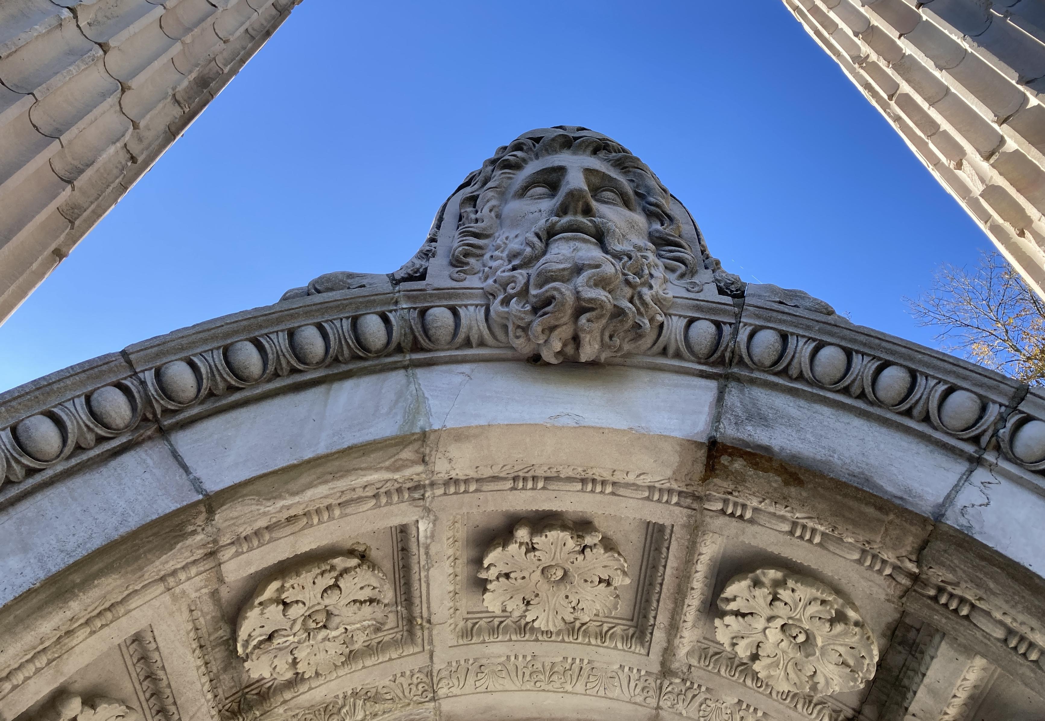 Looking up at a marble arch. A bearded head sits at the top. Beautiful flowers and edgework are carved into the marble. On both sides, we can see marble columns rising into the blue sky.