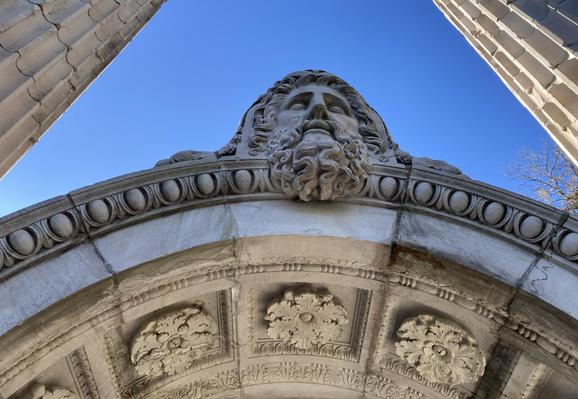 Looking up at a marble arch. A bearded head sits at the top. Beautiful flowers and edgework are carved into the marble. On both sides, we can see marble columns rising into the blue sky. 