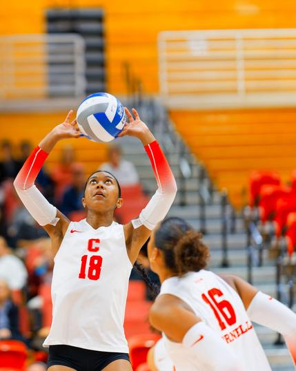 On the left half of the frame a woman has a volleyball in her hands stretched abover her head,  she's wearing a white tank top with C 18 in RED and has arm warmers that are white on top going to red on a gradient at the bottom,  no 16 similarly attired is approaching from her front and we can only see her back,  behind the two of them are partially full bleachers
