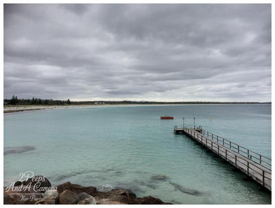 A wide angle landscape photo of a calm turquoise coloured ocean with a long wooden jetty extending from the right foreground.

The jetty leads out toward a small red object floating in the water. The beach runs along the left side of the frame, backed by low coastal scrub and houses under a heavily clouded, grey sky.