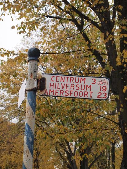 Fietswegwijzer: Centrum 3, Hilversum 19, Amersfoort 23. Op de achtergrond bomen met geel blad.