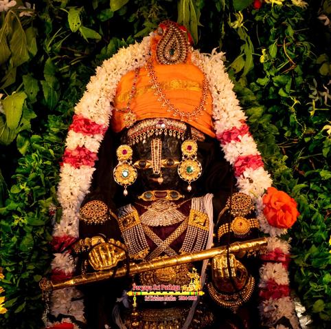 A very closeup of idol of Hindu deity Krishna in red ("grape red"), visible only waist above; wearing gold palm coverings, an orange turban with a head ornament that looks like a peacock feather, and lots of gold ornaments; holding a gilded flute between hands. A thick string of green holy basil leaves and a string of white flowers (some red roses) around Krishna. A dense arrangement of green holy basil leaves behind Krishna.